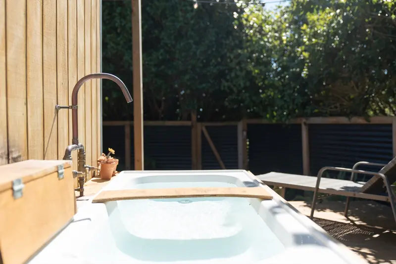 An outdoor bath beside a timber wall, with a bath tray and leafy garden beyond